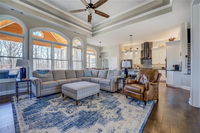 living room with dark wood finished floors, a tray ceiling, ornamental molding, ceiling fan with notable chandelier, and a wealth of natural light