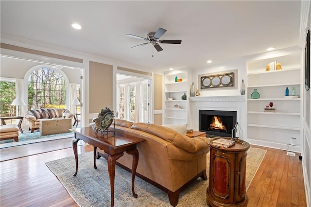 living area featuring hardwood / wood-style floors, built in features, recessed lighting, ornamental molding, and a lit fireplace