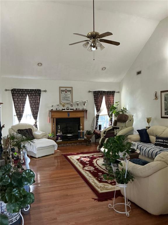 living room with high vaulted ceiling, a fireplace with raised hearth, visible vents, ceiling fan, and wood finished floors