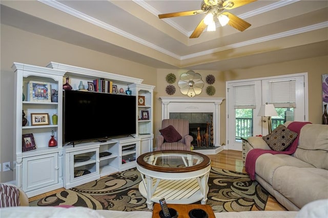 living area with ceiling fan, wood finished floors, ornamental molding, a tiled fireplace, and a raised ceiling