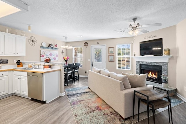 living room featuring sink, light hardwood / wood-style flooring, a stone fireplace, a textured ceiling, and ceiling fan with notable chandelier