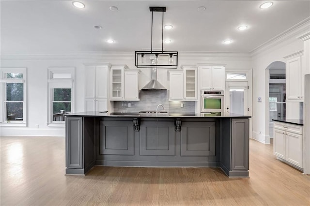 kitchen featuring arched walkways, ornamental molding, white cabinets, dark countertops, and wall chimney exhaust hood