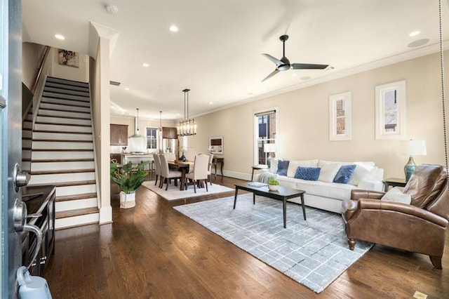 living area featuring recessed lighting, ceiling fan with notable chandelier, dark wood-style flooring, ornamental molding, and stairway