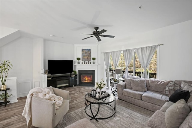 living room featuring a decorative wall, a ceiling fan, a warm lit fireplace, a wainscoted wall, and wood finished floors