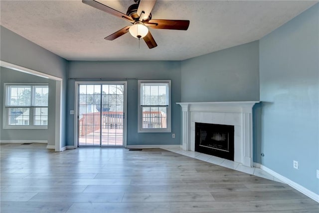 unfurnished living room with a tiled fireplace, light wood-style floors, a ceiling fan, a textured ceiling, and baseboards