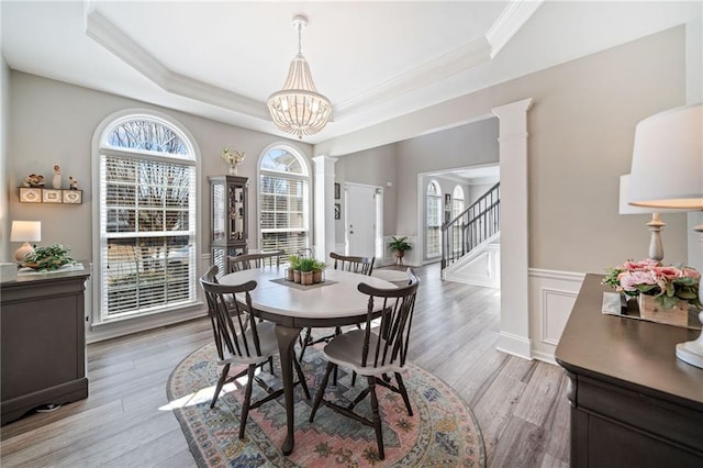 dining space featuring a wainscoted wall, light wood finished floors, ornate columns, ornamental molding, and a raised ceiling