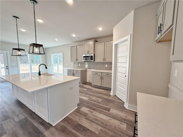 kitchen featuring stainless steel microwave, decorative backsplash, a sink, an island with sink, and wood finished floors