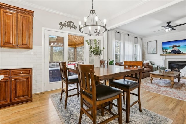 dining space with light wood finished floors, a glass covered fireplace, crown molding, and ceiling fan with notable chandelier