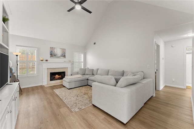 living room with baseboards, a ceiling fan, a fireplace with flush hearth, light wood-style floors, and high vaulted ceiling