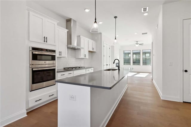 kitchen featuring wall chimney range hood, sink, a kitchen island with sink, hanging light fixtures, and white cabinets