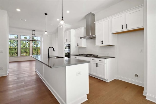 kitchen with wall chimney range hood, sink, white cabinetry, an island with sink, and decorative light fixtures