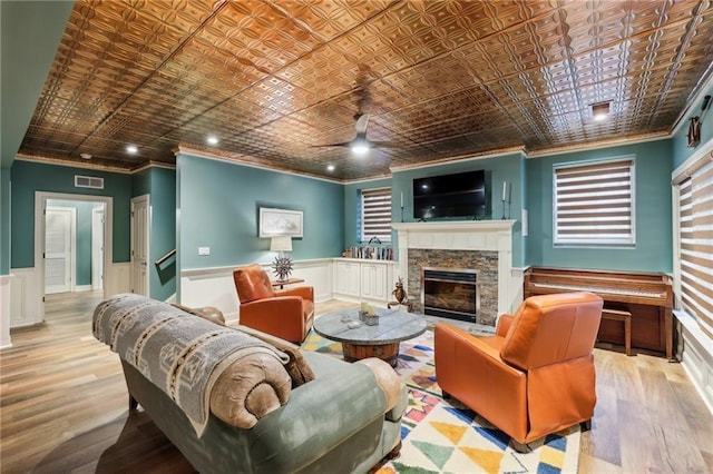 living room with crown molding, an ornate ceiling, a fireplace, a ceiling fan, and wainscoting
