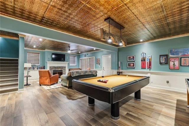 game room featuring an ornate ceiling, ornamental molding, wood finished floors, a stone fireplace, and a wainscoted wall