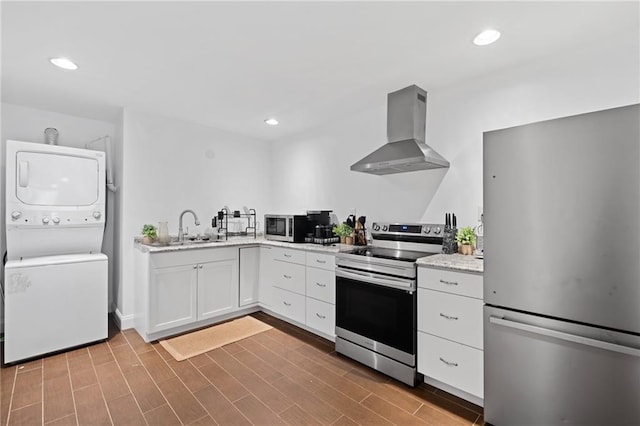 kitchen with stainless steel appliances, wall chimney exhaust hood, stacked washer / dryer, a sink, and white cabinets