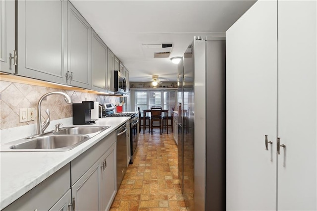 kitchen featuring a sink, gray cabinetry, ceiling fan, brick floor, and appliances with stainless steel finishes