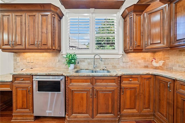 kitchen featuring tasteful backsplash, stainless steel dishwasher, brown cabinetry, a sink, and light stone countertops