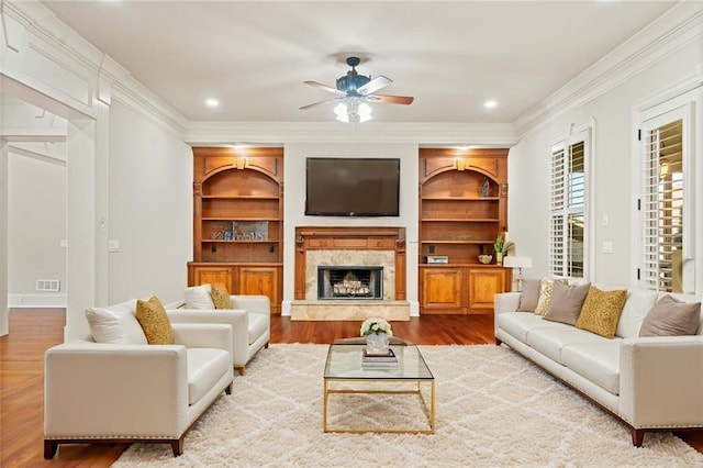 living area with built in features, crown molding, a premium fireplace, a ceiling fan, and wood finished floors