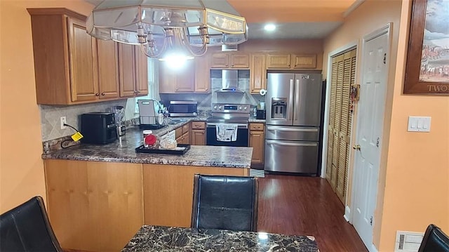 kitchen with stainless steel appliances, hanging light fixtures, wall chimney exhaust hood, dark countertops, and dark wood finished floors