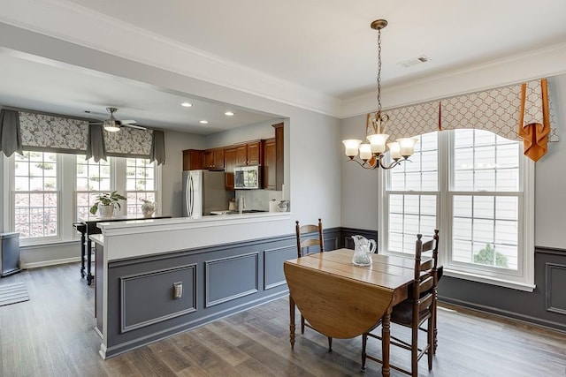 dining room featuring wood finished floors, ceiling fan with notable chandelier, a wainscoted wall, visible vents, and ornamental molding