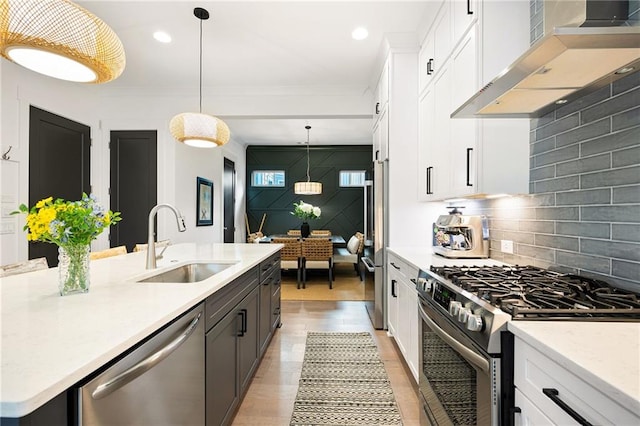 kitchen with appliances with stainless steel finishes, wall chimney exhaust hood, white cabinets, decorative light fixtures, and sink