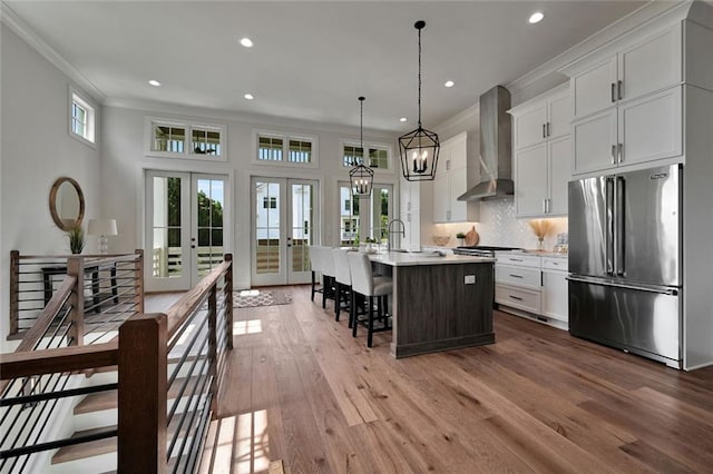 kitchen with high quality fridge, white cabinetry, a kitchen island with sink, wall chimney range hood, and french doors