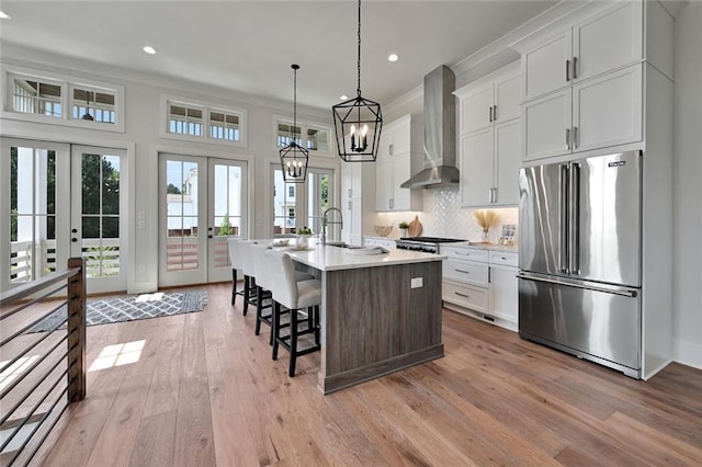 kitchen featuring white cabinets, french doors, stainless steel appliances, a center island with sink, and wall chimney exhaust hood