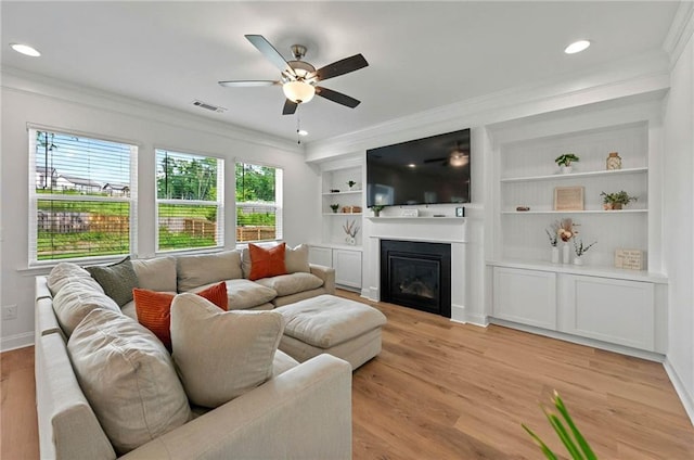 living area with crown molding, ceiling fan, a glass covered fireplace, light wood finished floors, and baseboards