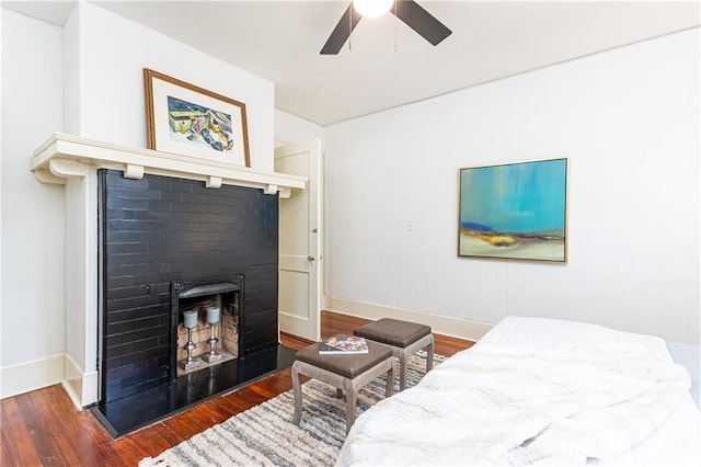 bedroom featuring baseboards, a fireplace with raised hearth, wood finished floors, and a ceiling fan