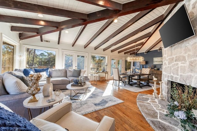 living room with hardwood / wood-style floors, a notable chandelier, lofted ceiling with beams, a stone fireplace, and french doors