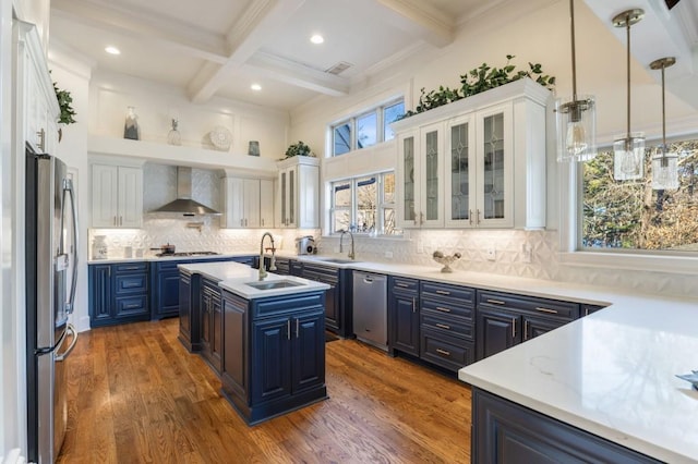 kitchen featuring wall chimney range hood, a kitchen island with sink, stainless steel appliances, white cabinets, and blue cabinets