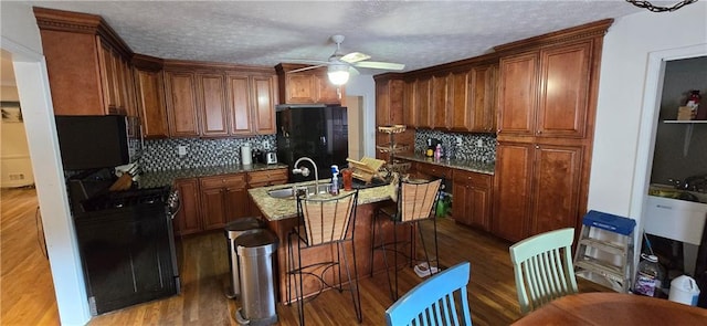 kitchen featuring dark wood-style flooring, a sink, ceiling fan, an island with sink, and black appliances