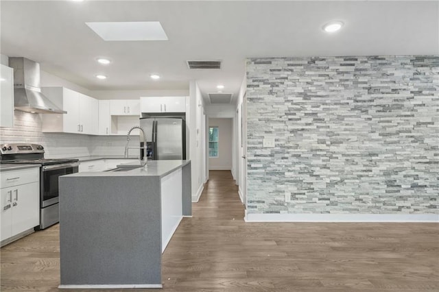 kitchen featuring wood-type flooring, appliances with stainless steel finishes, a kitchen island with sink, white cabinetry, and wall chimney range hood