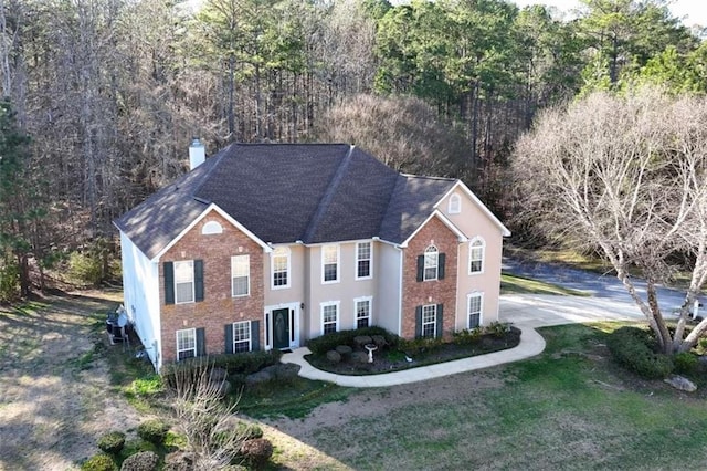 colonial-style house featuring stucco siding, brick siding, a forest view, and a chimney