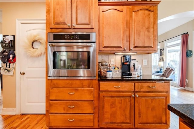 kitchen featuring stainless steel oven, dark stone countertops, backsplash, and light hardwood / wood-style flooring