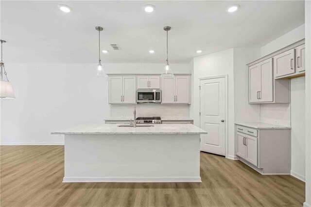 kitchen with light wood-type flooring, decorative light fixtures, light stone counters, and an island with sink