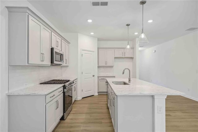 kitchen featuring light stone countertops, hanging light fixtures, a center island with sink, appliances with stainless steel finishes, and light wood-type flooring