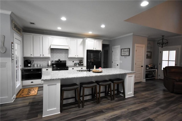 kitchen with custom exhaust hood, a kitchen island with sink, range with electric stovetop, white cabinetry, and black refrigerator with ice dispenser