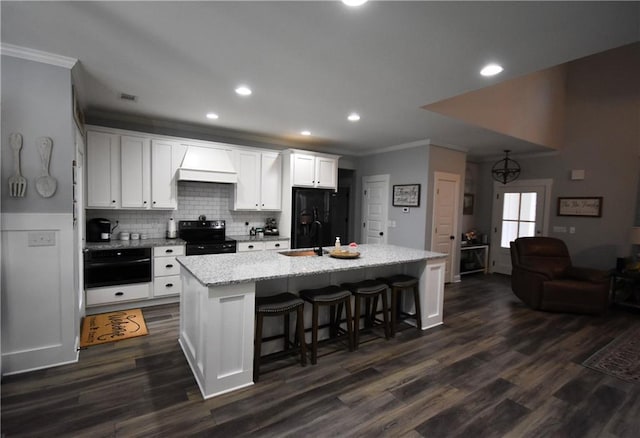 kitchen with a kitchen island with sink, custom exhaust hood, white cabinets, black appliances, and a sink