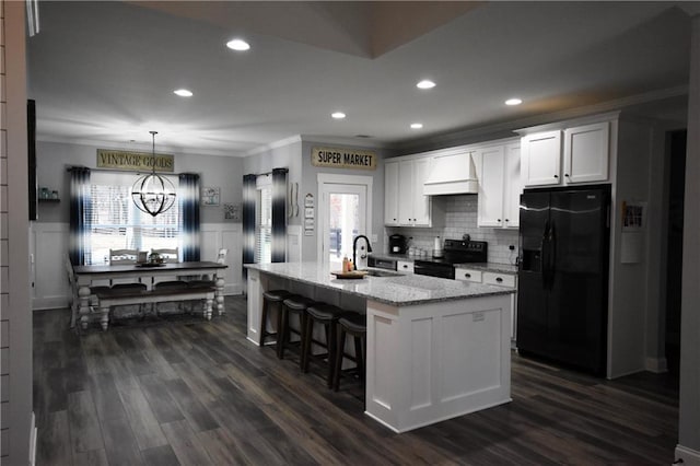 kitchen with premium range hood, black appliances, a sink, white cabinets, and wainscoting