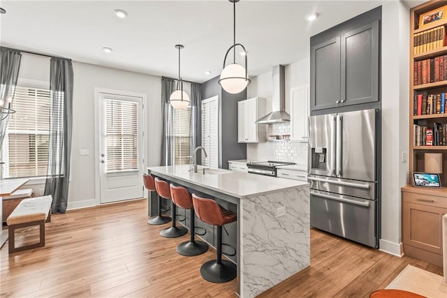 kitchen with stainless steel appliances, a kitchen island with sink, sink, wall chimney range hood, and hanging light fixtures