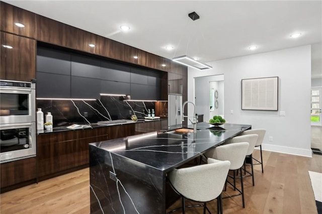 kitchen with light hardwood / wood-style floors, a kitchen island with sink, stainless steel double oven, backsplash, and a kitchen bar