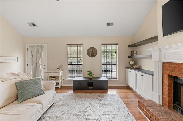 living room with a brick fireplace, visible vents, vaulted ceiling, and light wood-style flooring