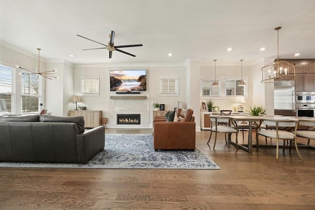 living room with a chandelier, crown molding, a warm lit fireplace, wood finished floors, and ceiling fan