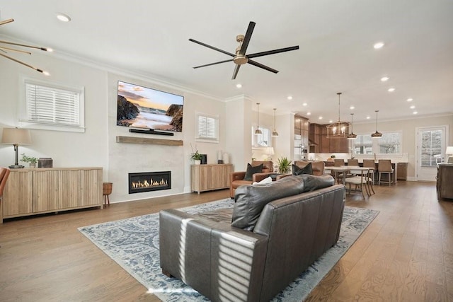 living area featuring ornamental molding, a ceiling fan, a warm lit fireplace, wood finished floors, and recessed lighting
