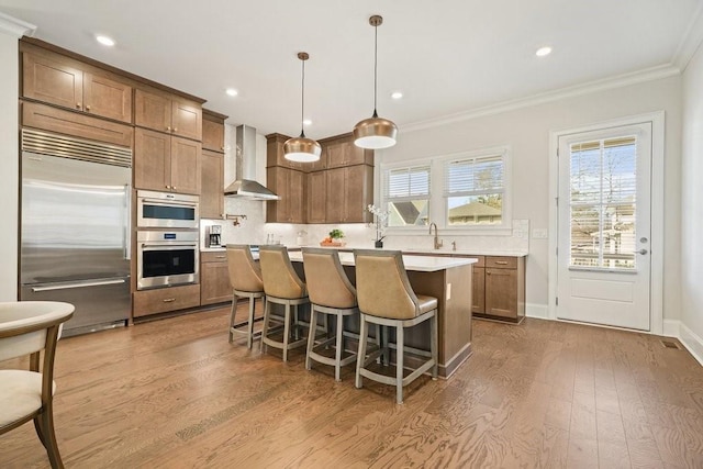 kitchen with appliances with stainless steel finishes, wall chimney exhaust hood, wood finished floors, ornamental molding, and light countertops