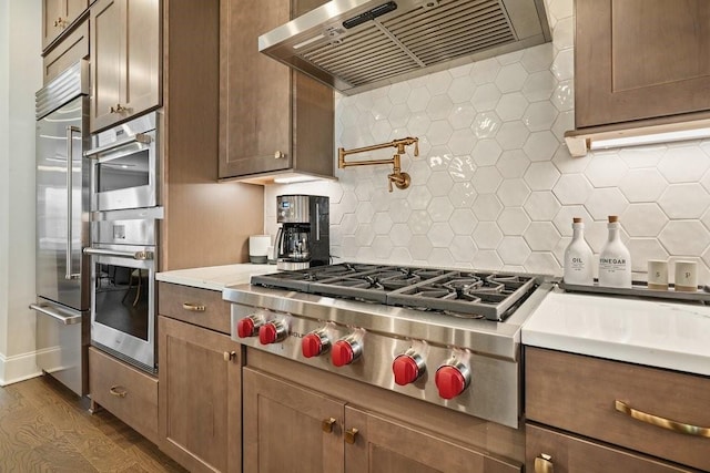 kitchen with appliances with stainless steel finishes, exhaust hood, dark wood finished floors, backsplash, and light countertops