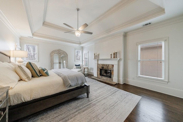 bedroom with visible vents, a raised ceiling, dark wood finished floors, crown molding, and a stone fireplace