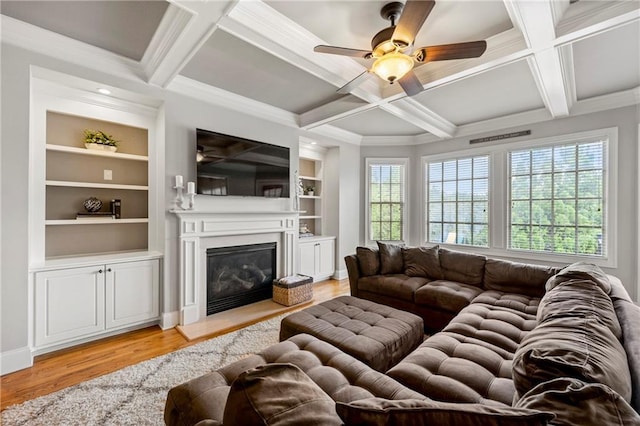 living area featuring light wood-style floors, a glass covered fireplace, a ceiling fan, beamed ceiling, and coffered ceiling