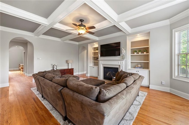 living area featuring crown molding, light wood finished floors, arched walkways, a glass covered fireplace, and a ceiling fan