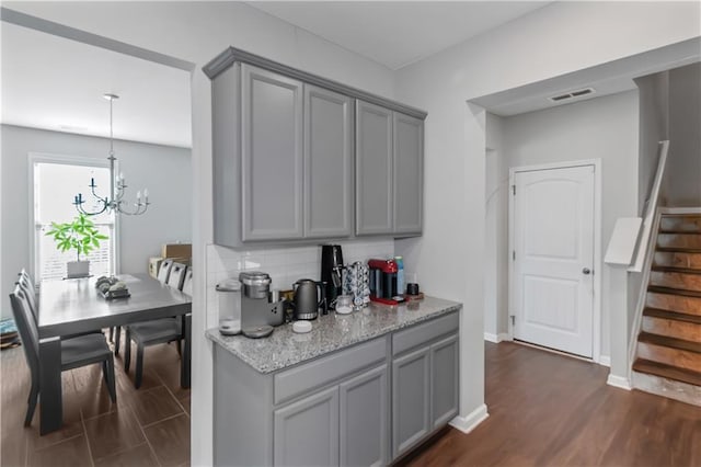 kitchen with light stone counters, decorative backsplash, gray cabinets, dark wood-style floors, and a notable chandelier